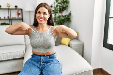 Genç esmer kadın evdeki kanepede oturuyor. Kendine güveni tam görünüyor. Kendini gururlu ve mutlu bir şekilde gösteriyor.. 