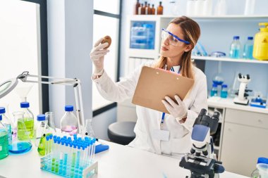 Young blonde woman wearing scientist uniform holding kiwi and clipboard at laboratory