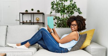 Genç Afro-Amerikan kadın gülümseyerek evde kendinden emin bir şekilde kitap okuyor.