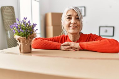 Orta yaşlı, kır saçlı, gülümseyen, kendine güvenen bir kadın yeni evinde pakete yaslanıyor.
