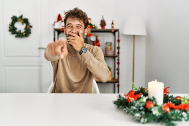 Genç, sakallı yakışıklı adam Noel süslemesi masasında oturmuş sana gülüyor, eli ağzında kamerayı işaret ediyor, utanç ifadesi takınıyor. 