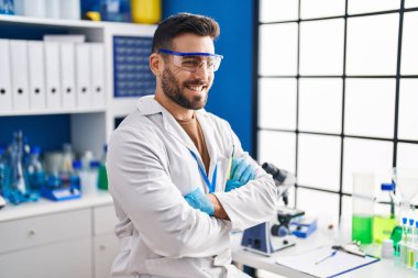 Laboratuvarda kollarını kavuşturup oturan bilim adamı üniforması giyen İspanyol genç adam.