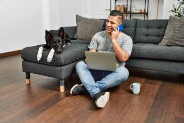 Genç İspanyol adam dizüstü bilgisayar kullanıyor ve akıllı telefonla konuşuyor evde köpekle yerde oturuyor..