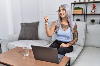 Middle age grey-haired woman using laptop at home smiling with happy face looking and pointing to the side with thumb up. 