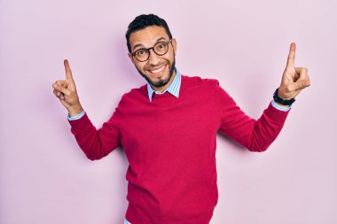 Hispanic man with beard wearing business shirt and glasses smiling amazed and surprised and pointing up with fingers and raised arms. 