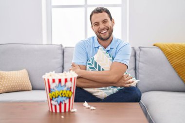 Hispanic man watching tv at home sitting on the sofa smiling and laughing hard out loud because funny crazy joke. 
