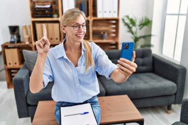 Beautiful woman doing online therapy session at consultation office smiling with an idea or question pointing finger with happy face, number one 
