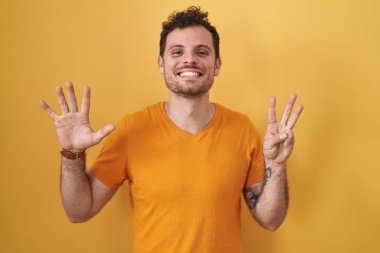 Sarı arka planda dikilen İspanyol genç adam sekiz numaralı parmaklarıyla gülümseyerek kendinden emin ve mutlu bir şekilde işaret ediyor.. 