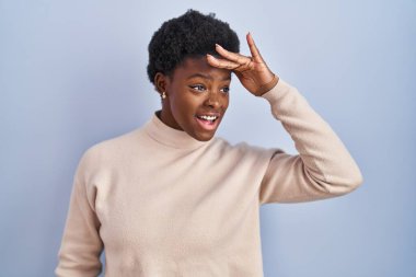 African american woman standing over blue background very happy and smiling looking far away with hand over head. searching concept. 