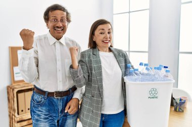 Middle age interracial couple holding recycling bin with plastic bottles at the office screaming proud, celebrating victory and success very excited with raised arms 