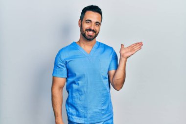 Yakışıklı, sakallı, mavi erkek hemşire üniforması giyen, neşeli bir şekilde gülümseyen ve avuç içi kameraya bakan bir İspanyol.. 