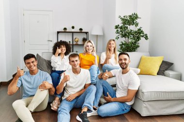 Group of people sitting on the sofa and floor at home smiling happy and positive, thumb up doing excellent and approval sign 