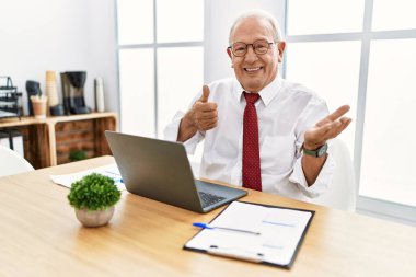 Ofiste çalışan kıdemli bir adam bilgisayarla avuç içini gösteriyor ve baş parmaklarıyla iyi bir hareket yapıyor, mutlu ve neşeli gülümsüyor. 