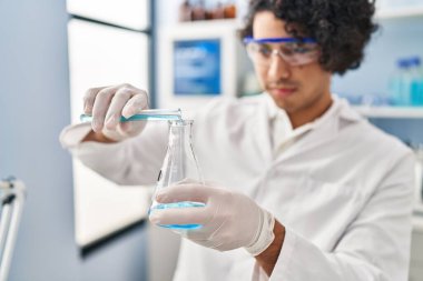 Laboratuvarda test tüpüne sıvı döken bilim adamı üniforması giyen İspanyol genç adam.