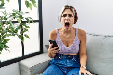 Young beautiful woman using smartphone typing message sitting on the sofa angry and mad screaming frustrated and furious, shouting with anger. rage and aggressive concept. 