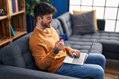Young hispanic man using laptop and credit card sitting on sofa at home