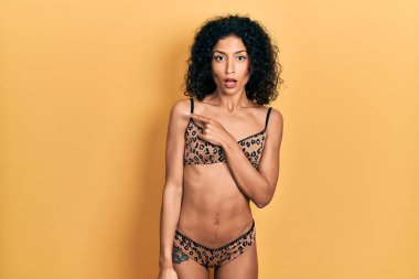 Young latin girl wearing bikini surprised pointing with finger to the side, open mouth amazed expression. 