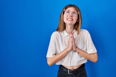 Beautiful woman standing over blue background begging and praying with hands together with hope expression on face very emotional and worried. begging. 