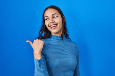 Mavi izole edilmiş arka planda duran genç Brezilyalı kadın mutlu bir yüzle gülümsüyor ve başparmağıyla yan tarafı işaret ediyor.. 