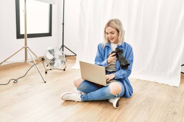 Fotoğrafçılık stüdyosunda çalışan güzel sarışın kadın yerde oturmuş bilgisayardaki fotoğrafları kontrol ediyor.
