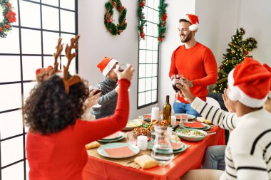 Mutlu bir şekilde gülümseyen bir grup genç Noel yemeği yiyor ve evde kadeh kaldırıyor..