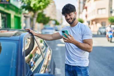 Young hispanic man using smartphone leaning on car at street