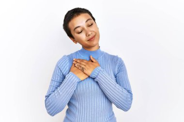 Kısa saçlı genç İspanyol kadın izole edilmiş arka planda duruyor elleri göğsünde, gözleri kapalı ve yüzünde minnet dolu bir jest ile gülümsüyor. sağlık kavramı. 
