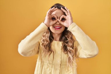 Sarı arka planda duran genç beyaz kadın dürbün gibi jest yapıyor. Dilini çıkarmış, gözleri parmaklarından bakıyor. çılgın ifade. 