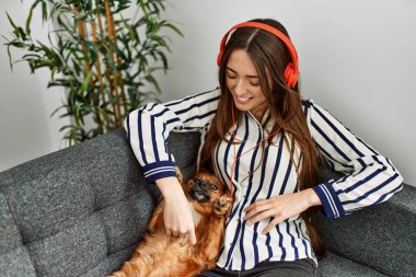 Young hispanic woman listening to music sitting on sofa with dog at home