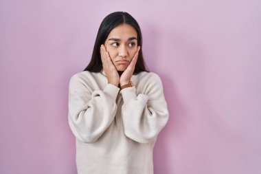 Pembe arka planda duran genç Güney Asyalı kadın yorgun eller yüzünü, depresyonu ve üzüntüyü örten, sorun yüzünden üzgün ve tedirgin 