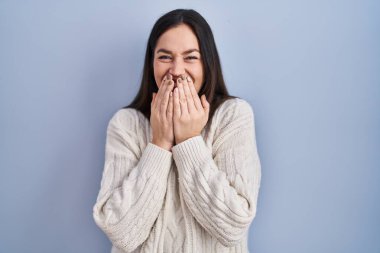 Genç esmer kadın mavi arka planda durup gülüyor ve utanıyor kıkırdıyor elleriyle ağzını kapatıyor, dedikodu ve skandal konsepti yapıyor. 