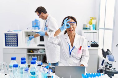 Laboratuvarda çalışan orta yaşlı bir kadın gülümseyen mutlu bir şekilde el pençe divan duruyor. 