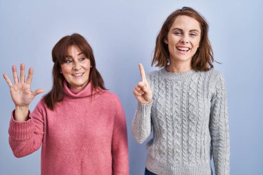 Anne ve kızı mavi arka planda dikiliyorlar ve altı numaralı parmaklarıyla işaret ediyorlar. Kendinden emin ve mutlu bir şekilde gülümsüyorlar.. 