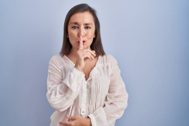 Middle age hispanic woman standing over blue background asking to be quiet with finger on lips. silence and secret concept. 