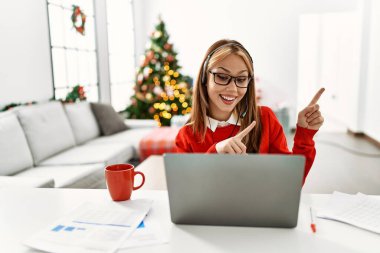 Young caucasian girl sitting on the table working using laptop by christmas tree smiling and looking at the camera pointing with two hands and fingers to the side. 