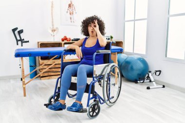 Young middle eastern woman sitting on wheelchair at physiotherapy clinic doing ok gesture shocked with surprised face, eye looking through fingers. unbelieving expression. 