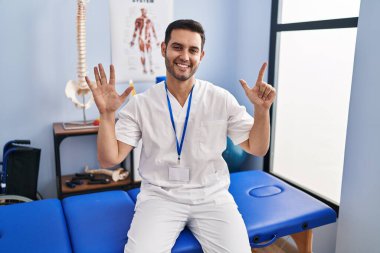 Sakallı ve sakallı genç bir adam ağrı kesici kliniğinde çalışıyor. Yedi numaralı parmaklarını gösterip işaret ediyor. Kendinden emin ve mutlu bir şekilde gülümsüyor.. 