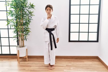 Karate kimonosu ve siyah kuşak giyen genç İspanyol kız dudaklarında parmaklarıyla sessiz olmak istiyor. sessizlik ve gizli konsept. 