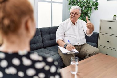 Senior psychologist man at consultation office approving doing positive gesture with hand, thumbs up smiling and happy for success. winner gesture. 
