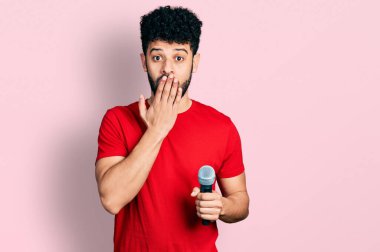 Sakallı genç Arap adam mikrofon kullanarak şarkı söylüyor. Ağzını eliyle kapatıyor, şok geçiriyor ve hata yapmaktan korkuyor. şaşırmış ifade 