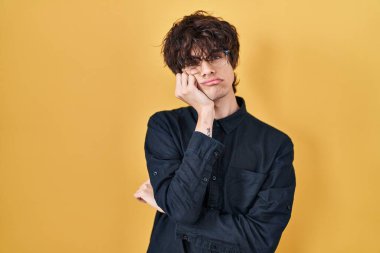 Young man wearing glasses over yellow background thinking looking tired and bored with depression problems with crossed arms. 