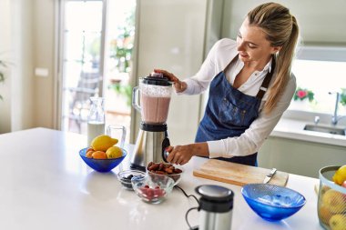 Genç sarışın kadın gülümsüyor. Kendine güveni tam. Smoothie 'yi sıvılaştırıyor.