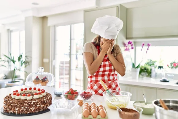 Beautiful young brunette pastry chef woman cooking pastries at the kitchen with sad expression covering face with hands while crying. depression concept.