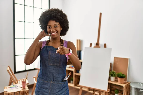 Young african american woman with afro hair at art studio smiling doing talking on the telephone gesture and pointing to you. call me.