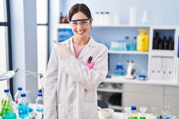 Young brunette woman working at scientist laboratory smiling cheerful pointing with hand and finger up to the side