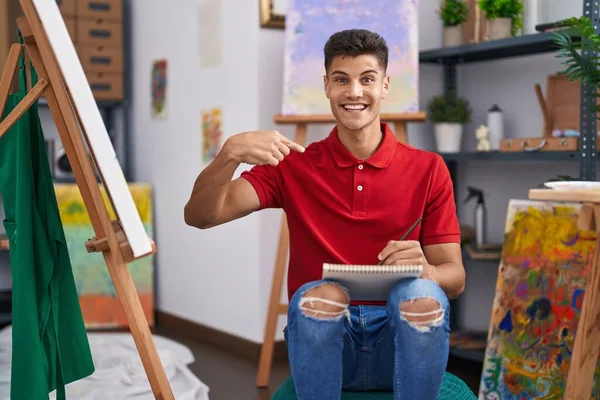 Young hispanic man drawing on art notebook pointing finger to one self smiling happy and proud