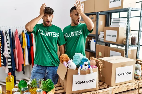 Young gay couple wearing volunteer t shirt at donations stand surprised with hand on head for mistake, remember error. forgot, bad memory concept.