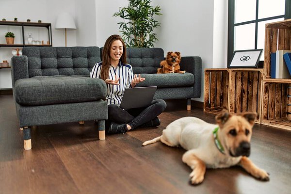 Young hispanic woman having video call sitting on floor with dogs at home