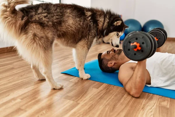 Young hispanic man smiling confident training using dumbbells with dog at sport center