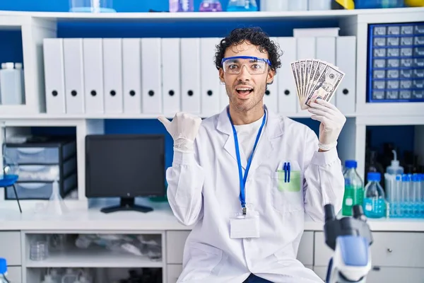Hispanic man working at scientist laboratory holding money pointing thumb up to the side smiling happy with open mouth